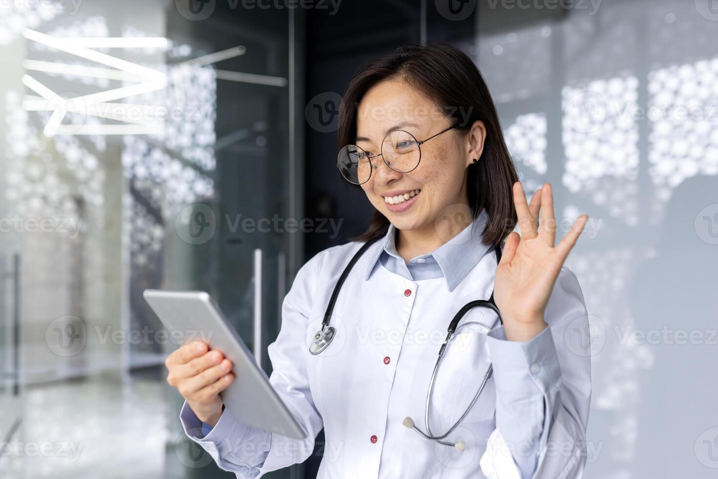 Cheerful and smiling asian female doctor talking to patient online remotely, woman standing in clinic and using tablet computer for call and online patient consultation. photo