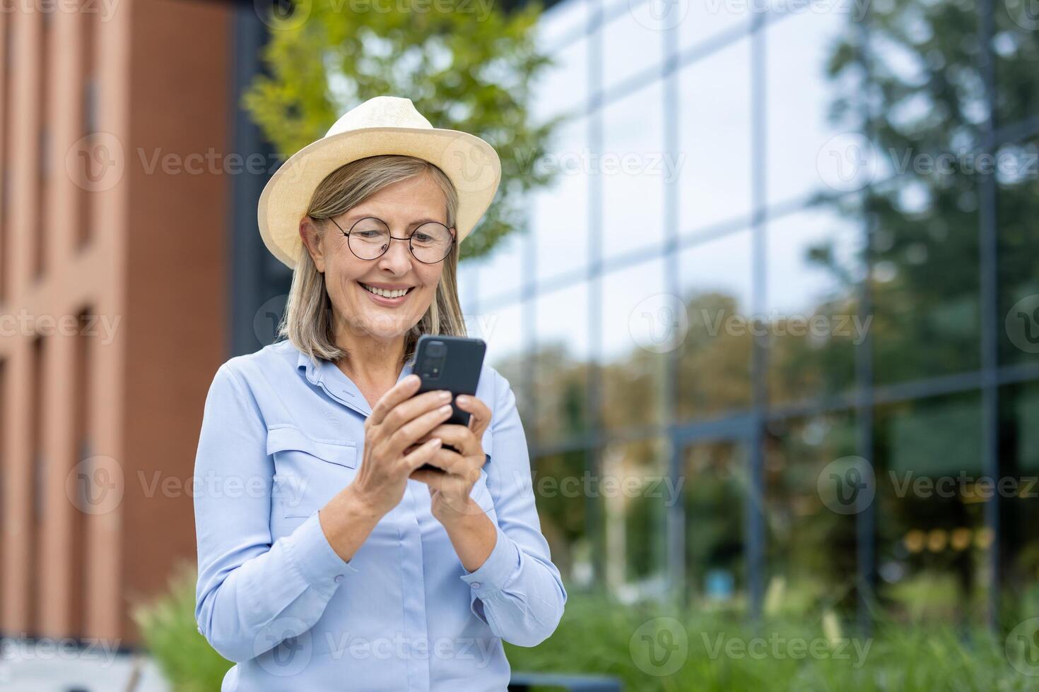 An older woman smiles while looking at her phone, standing outside with a building in the background. She is wearing a hat and glasses. photo
