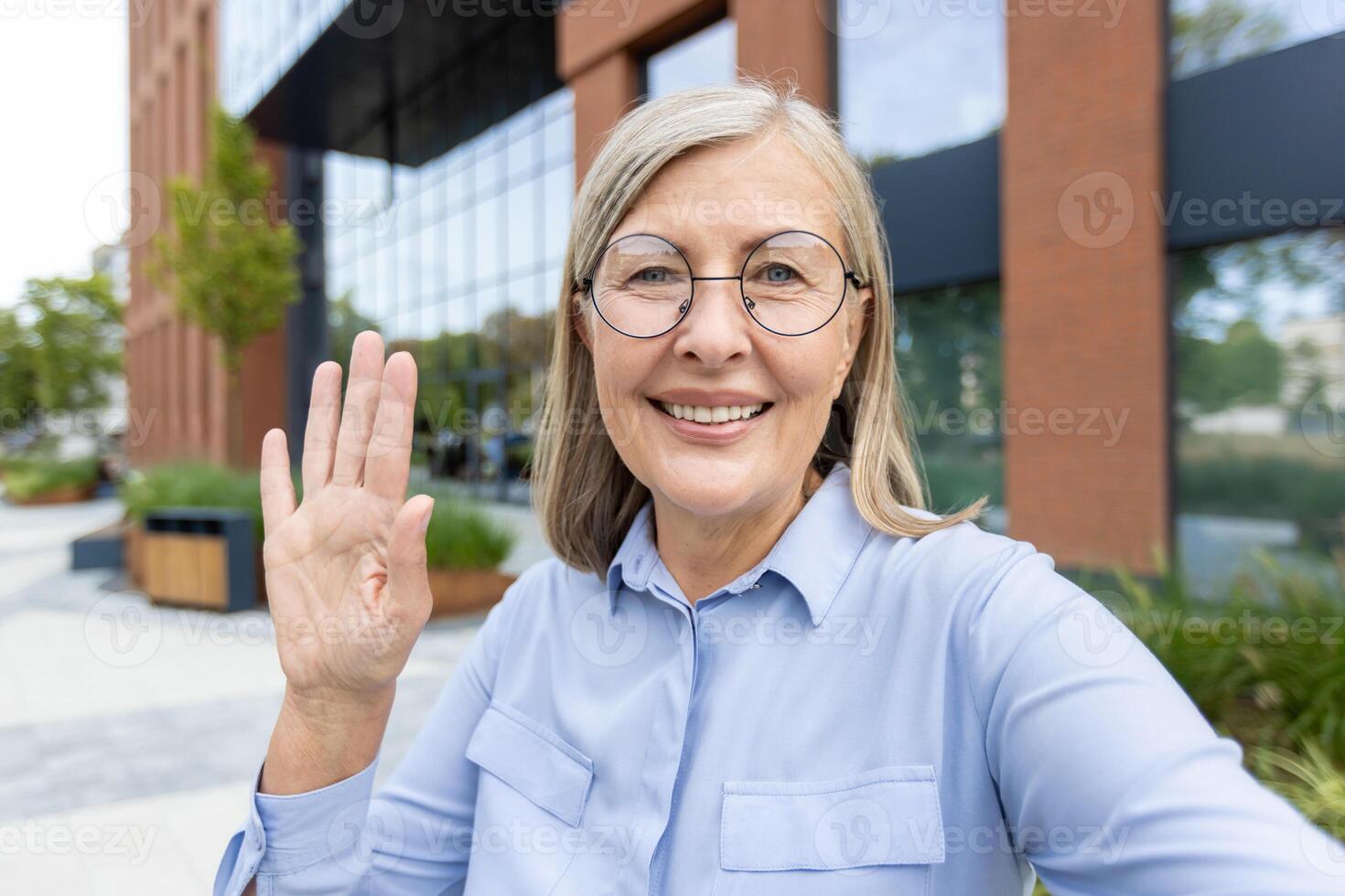 Smiling senior woman waves during a call, possibly using a phone, outdoors near a modern building photo