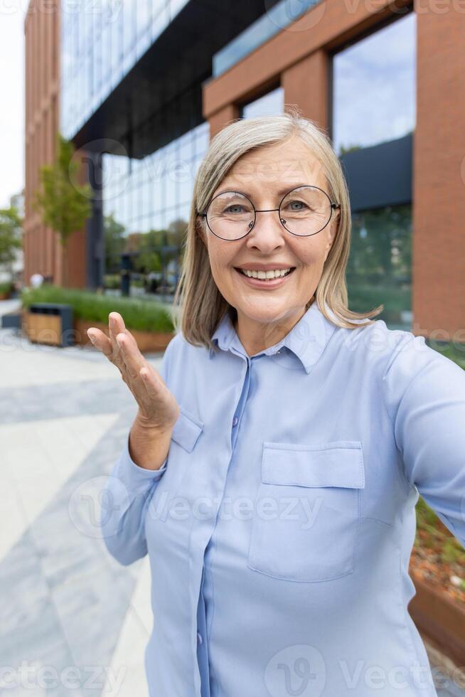 An attractive senior woman in glasses smiles and gestures while taking a selfie outside near a modern building. photo