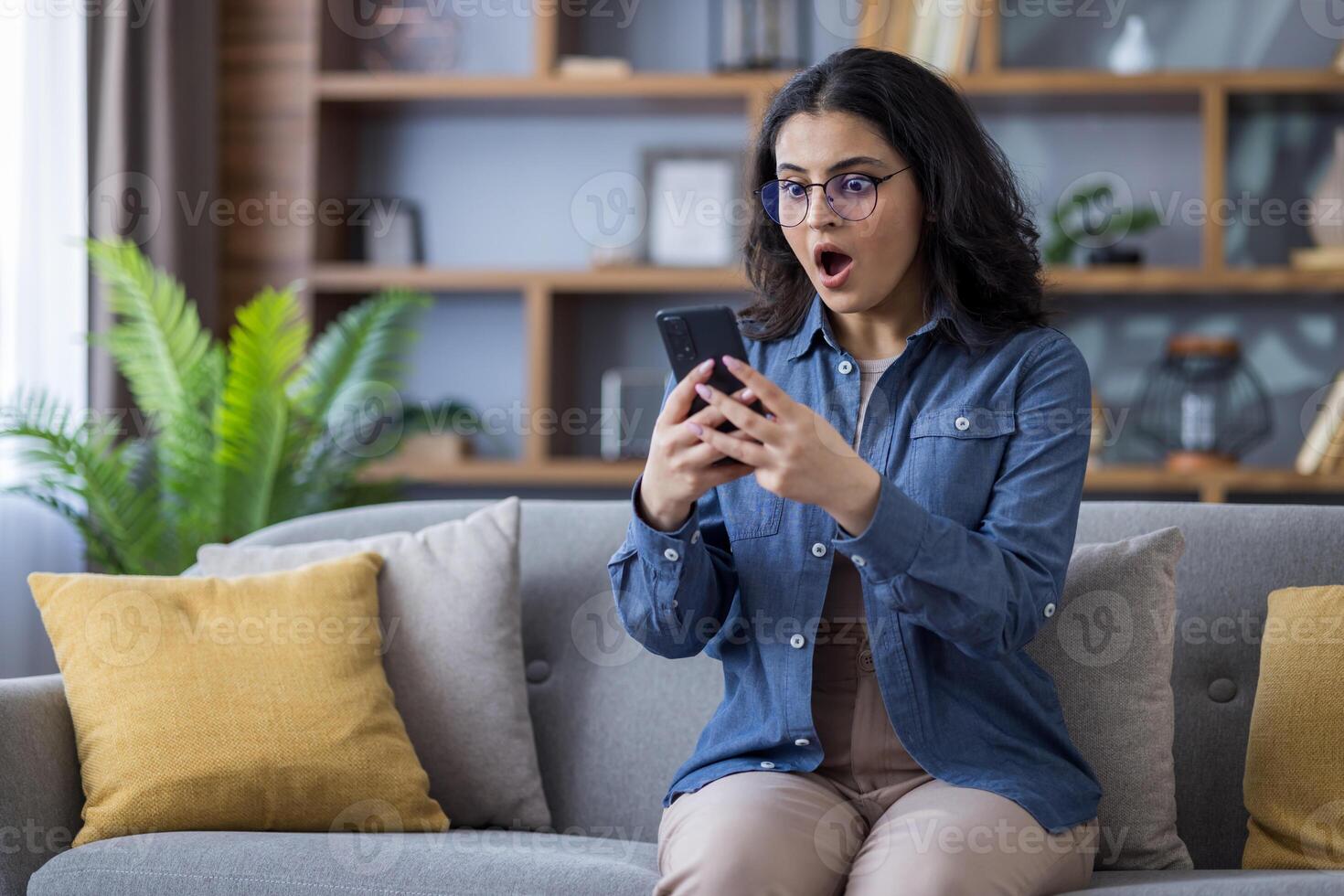 Photo of a surprised Indian woman sitting on the couch at home and looking shocked at the screen of the mobile phone she is holding.
