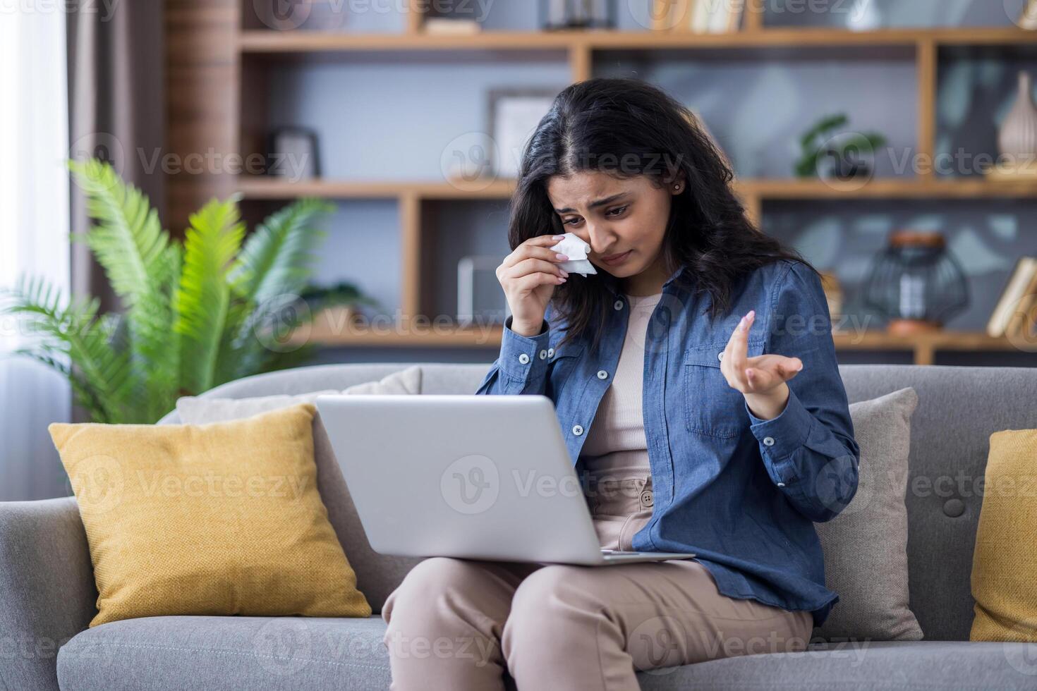 Upset young Indian woman sitting on sofa at home, holding laptop on lap, talking on call and crying, wiping tears with napkin. photo