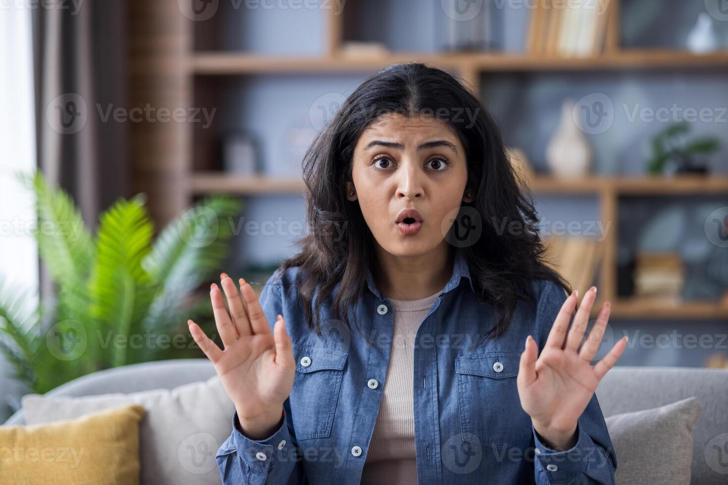 Close-up portrait of a young worried Indian woman sitting on the couch at home, looking at the camera and showing a stop sign with her hands. photo