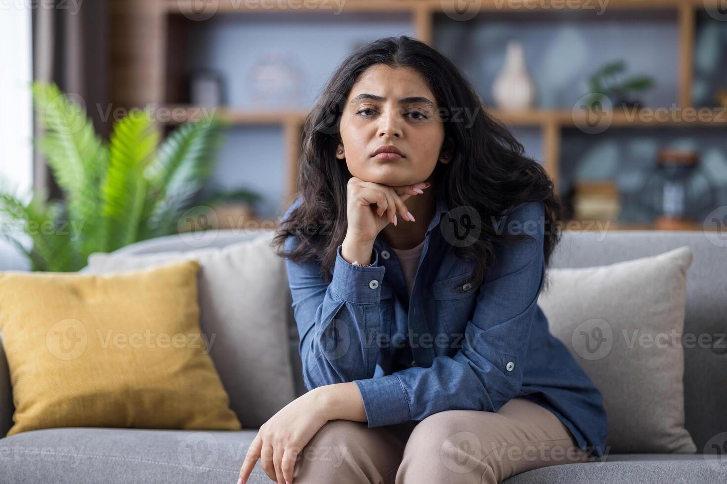 A woman looks directly at the camera while sitting on a couch, hands supporting her chin. She has a serious, thoughtful expression. photo