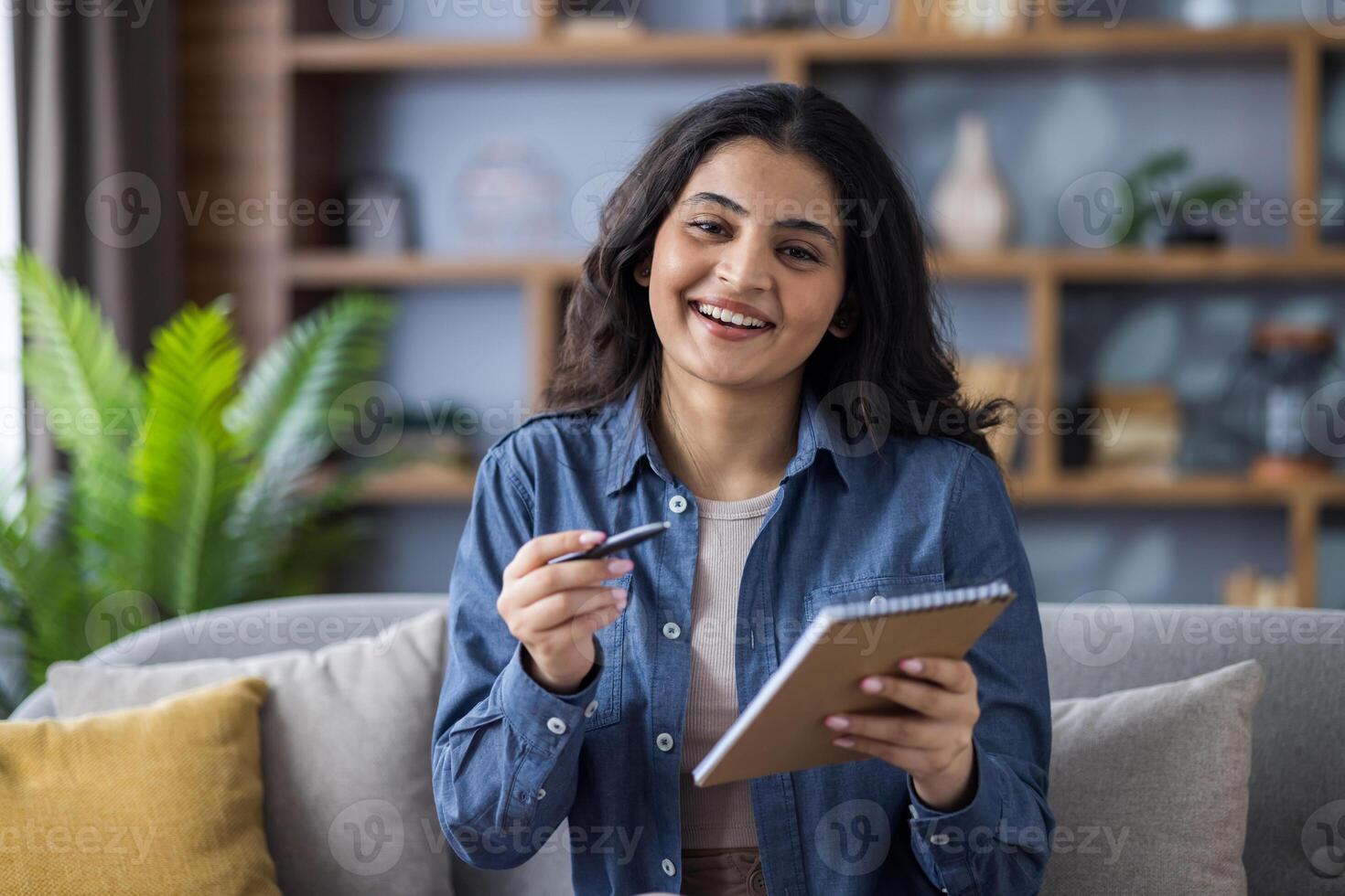 A smiling young woman holds a pen and notepad, possibly preparing to take notes or brainstorming ideas in a comfortable setting. photo