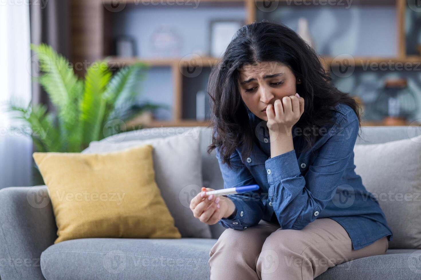 A woman looks anxiously at a pregnancy test, sitting on a couch in a living room. This image conveys uncertainty about the future. photo