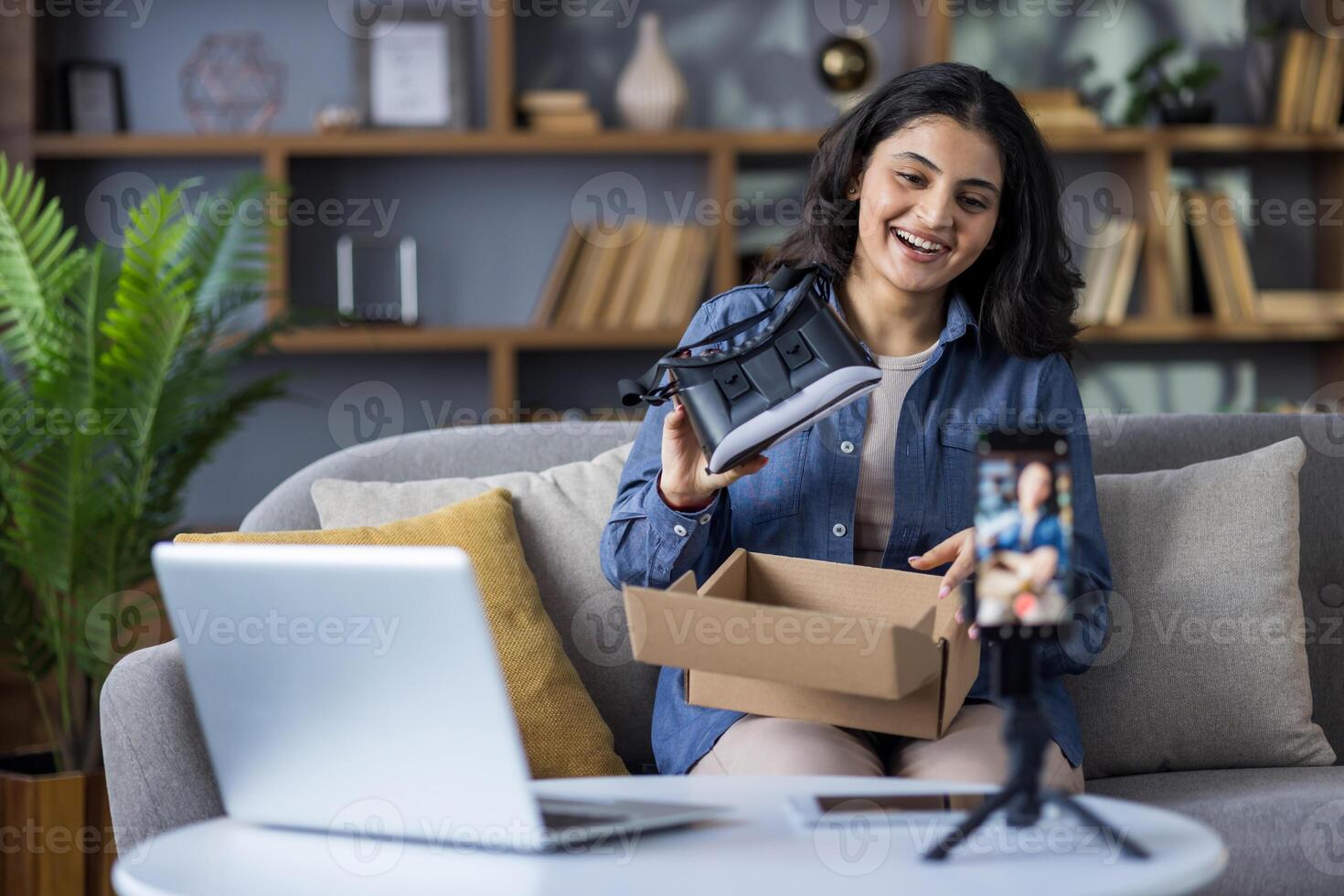 Smiling young Indian woman records a blog on her phone camera, conducts a broadcast and shows the unpacking of the purchased product, holds a box with virtual glasses. photo