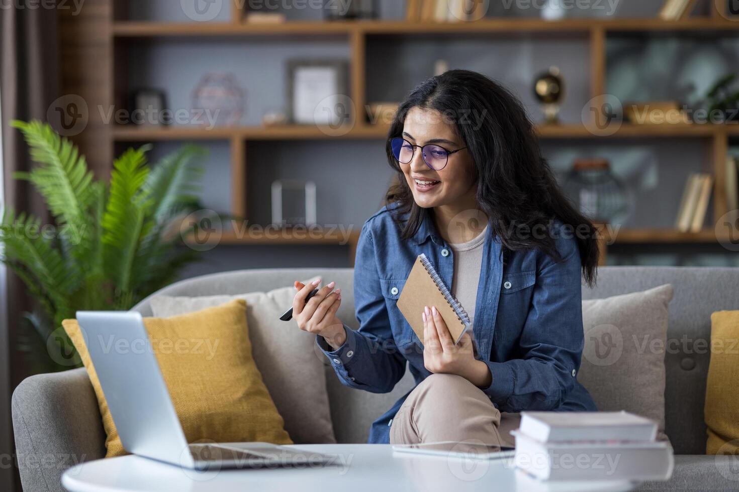 A smiling woman with glasses is sitting on a couch, appearing to be in a call on her laptop, holding a notebook. photo