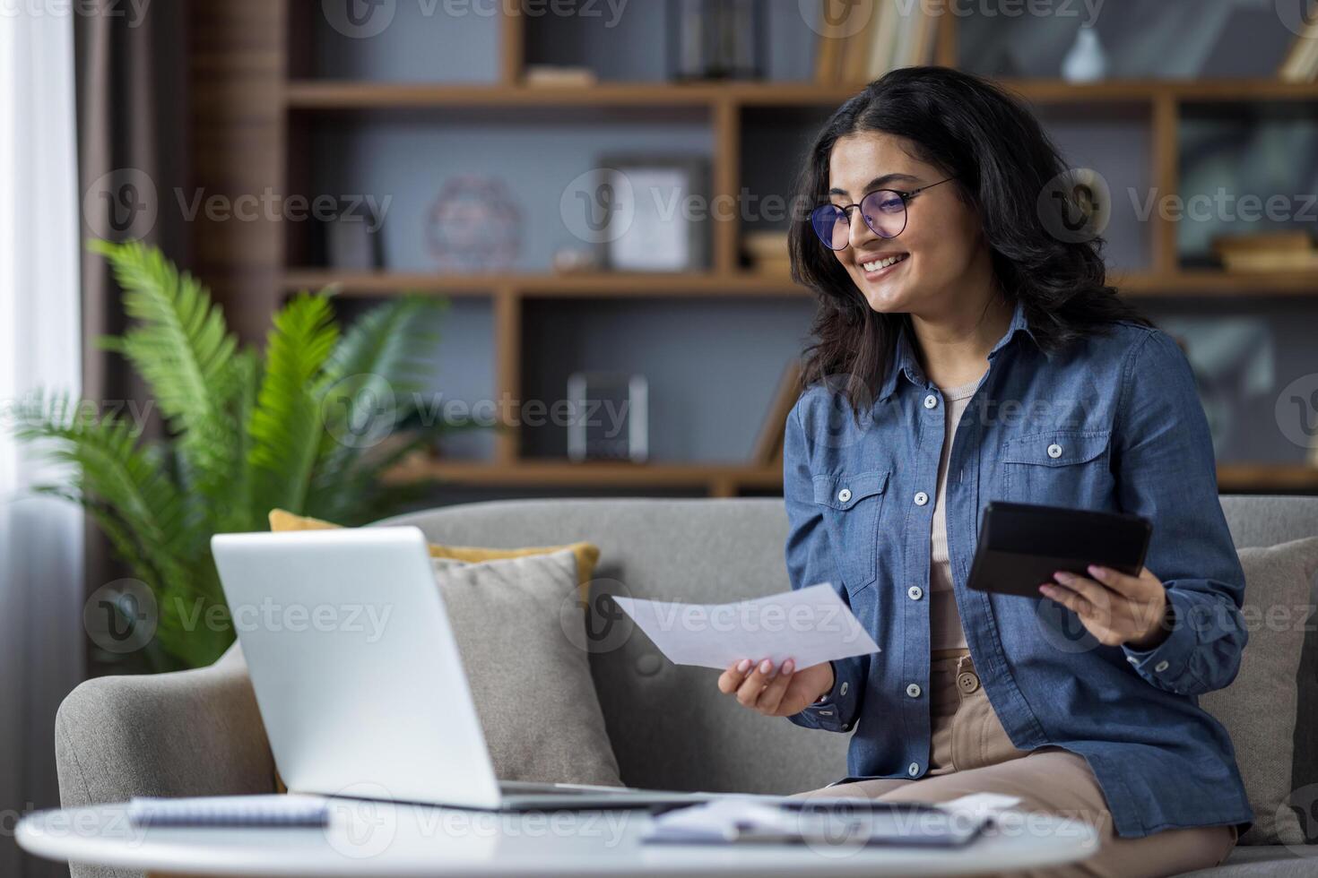 A smiling young woman works from home, holding documents and a tablet while using her laptop on the couch. photo