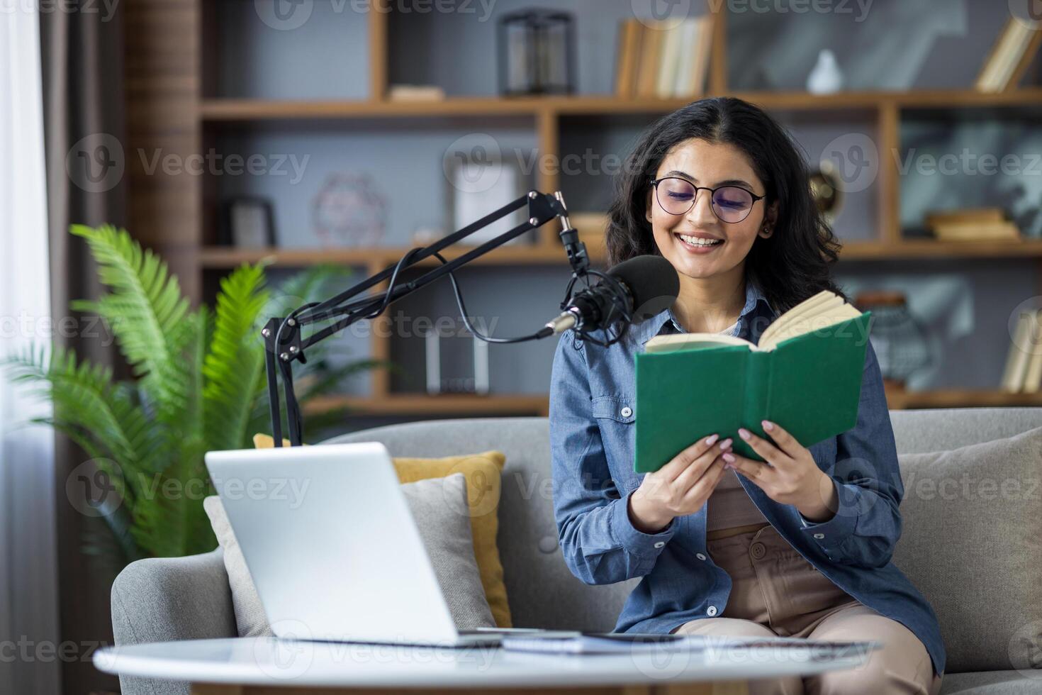 A cheerful woman in glasses enjoys a book during a podcast recording, sitting on a couch in a cozy setting, complete with a microphone and laptop. photo