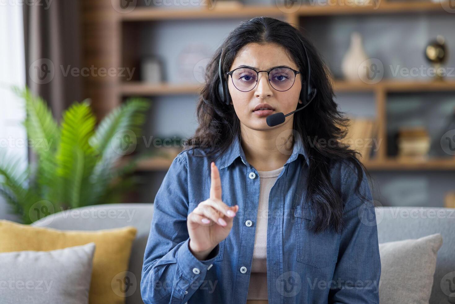Portrait of a young Indian woman wearing a headset at home, sitting on a sofa and talking to the camera, showing a stop gesture with her finger. photo