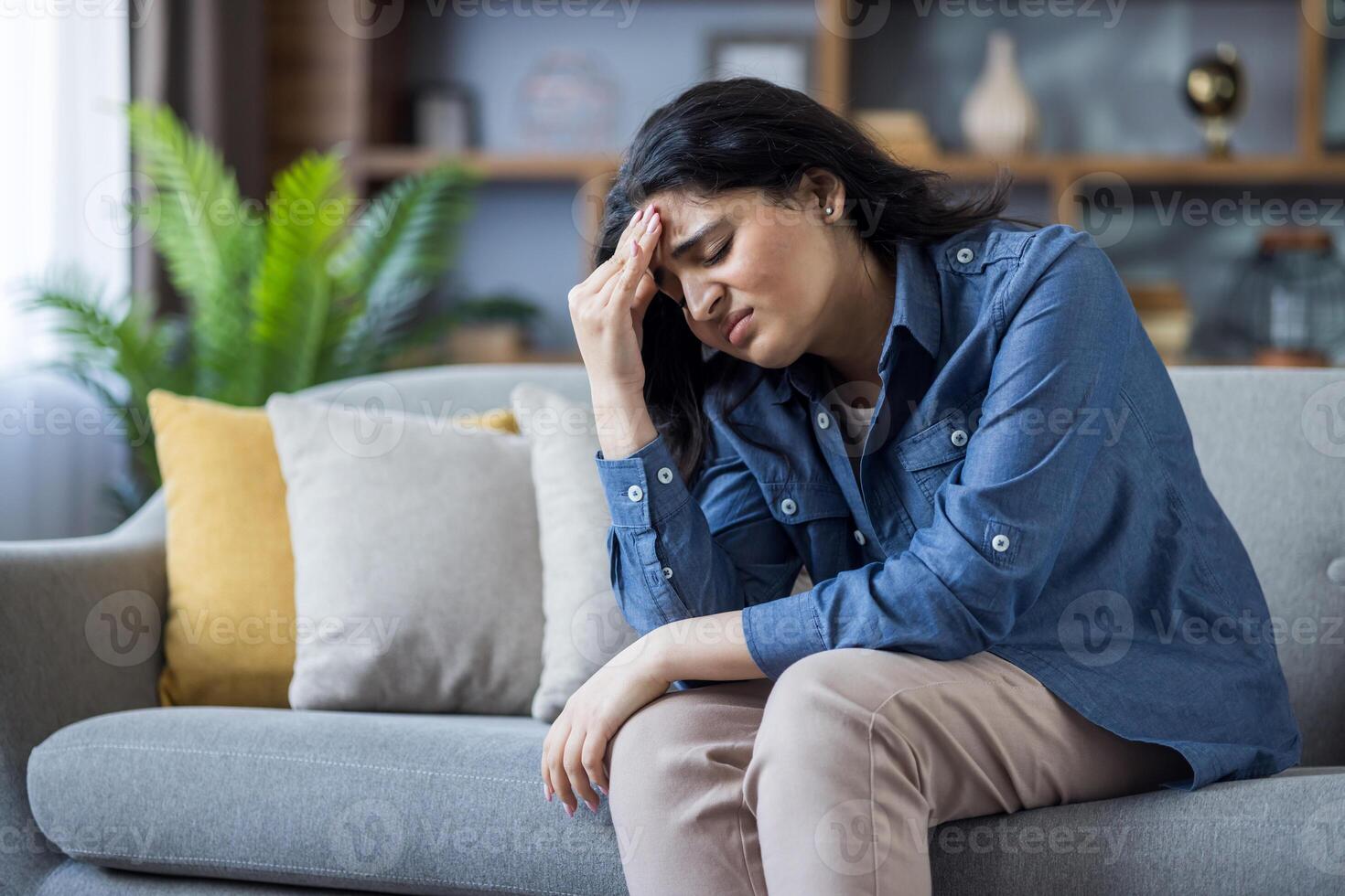 A young woman is experiencing a headache while sitting on a sofa at home, looking stressed. She is clutching her forehead. photo