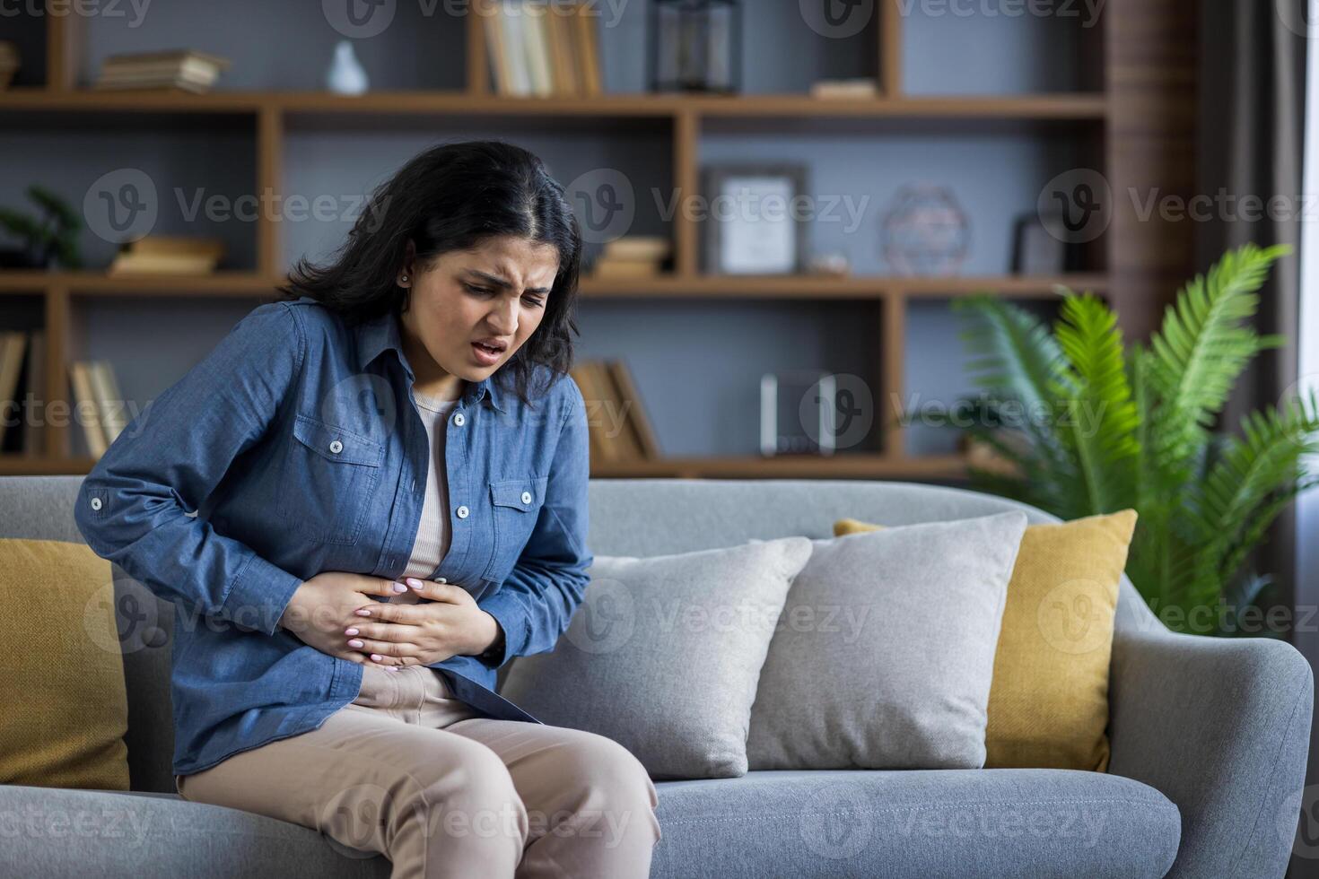 A woman, doubled over in pain, clutches her stomach while sitting on a sofa, indicating possible discomfort. photo