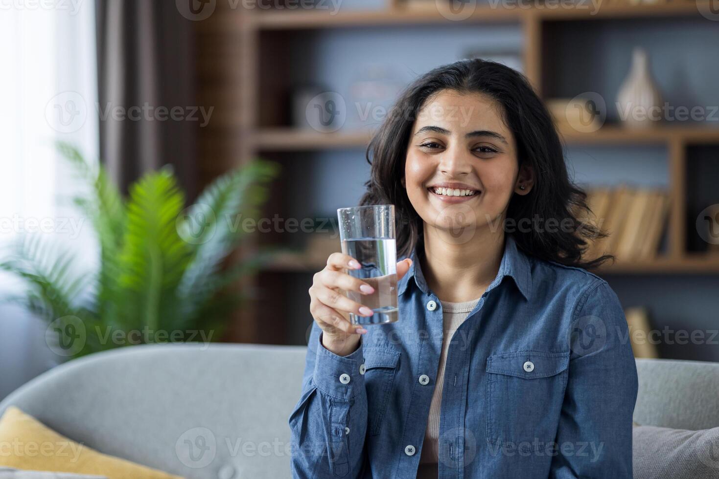 Close-up portrait of a young smiling Indian woman at home, sitting on the sofa, holding a glass of clean water in her hand, looking at the camera. photo