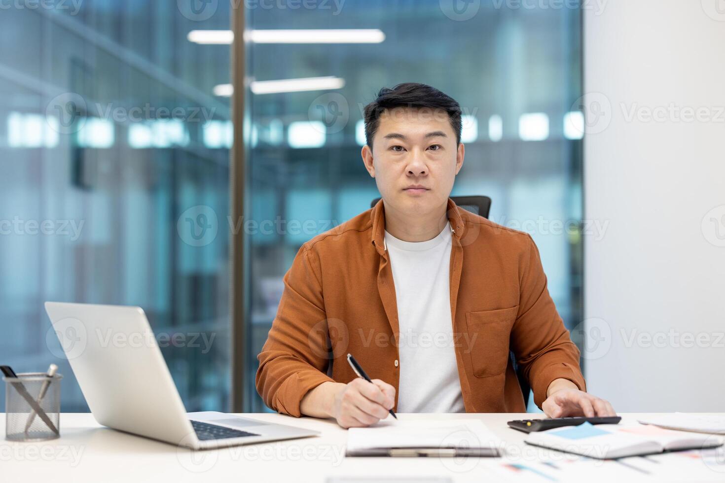 An Asian businessman in a modern office setting, working at his desk. He's focused, looking directly at the camera. photo