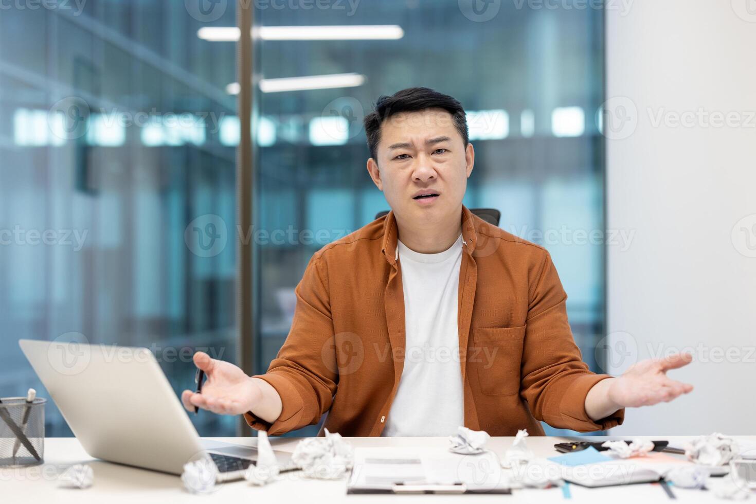 An Asian man, seemingly frustrated, sits at his desk with a laptop and crumpled paper, expressing confusion or stress. photo