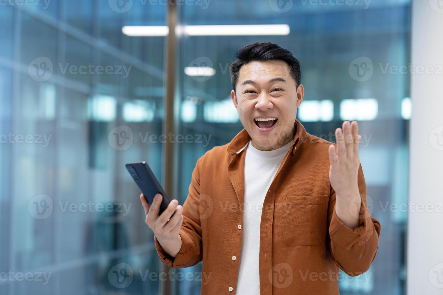 An Asian man, smiling broadly and gesturing with excitement while holding a smartphone, appears pleased with the content. photo