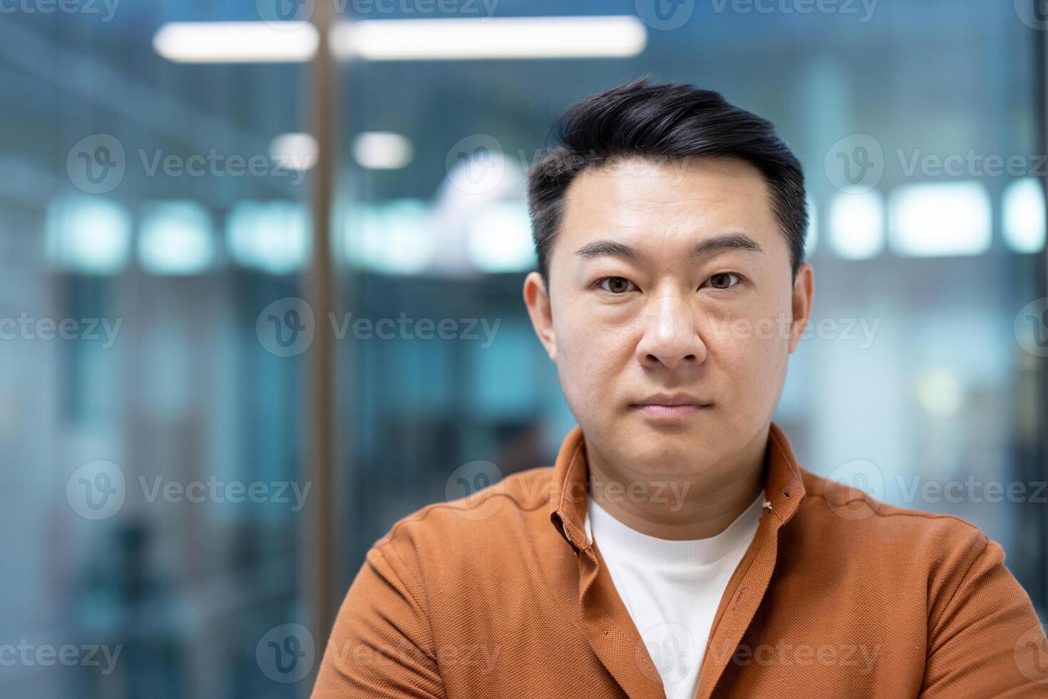 A portrait of an Asian man looking directly at the camera with a serious expression. He is wearing a brown shirt. photo