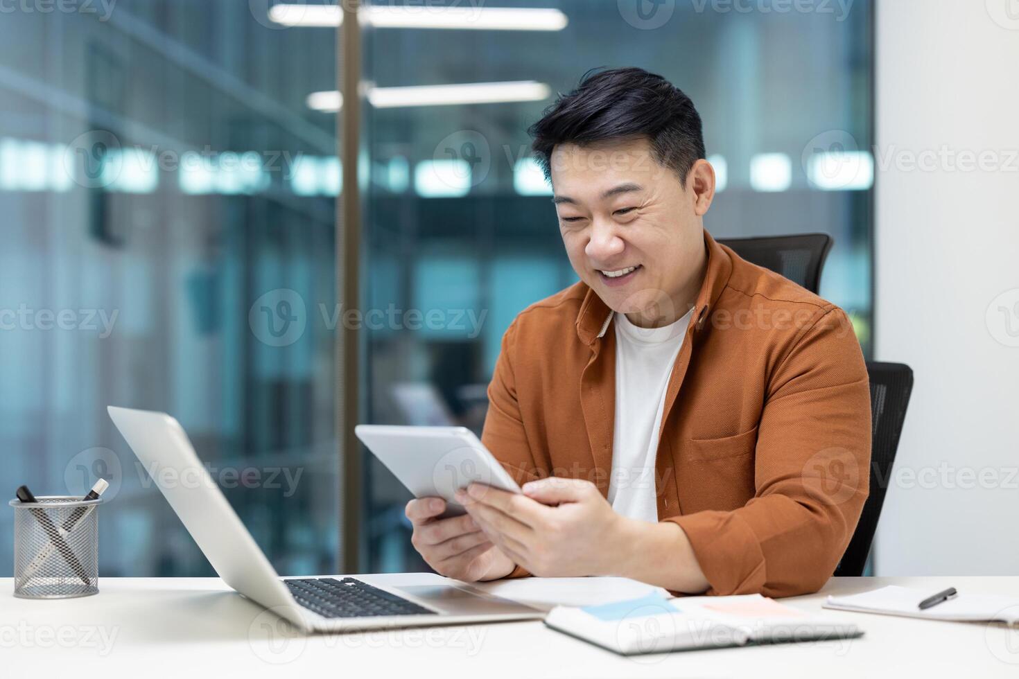 An Asian man smiles while working on a tablet and laptop at his desk in a modern office setting. photo
