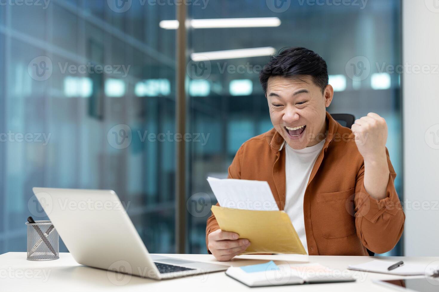 An excited Asian man celebrates with a raised fist after reading good news from a letter. He's sitting at a desk with a laptop. photo