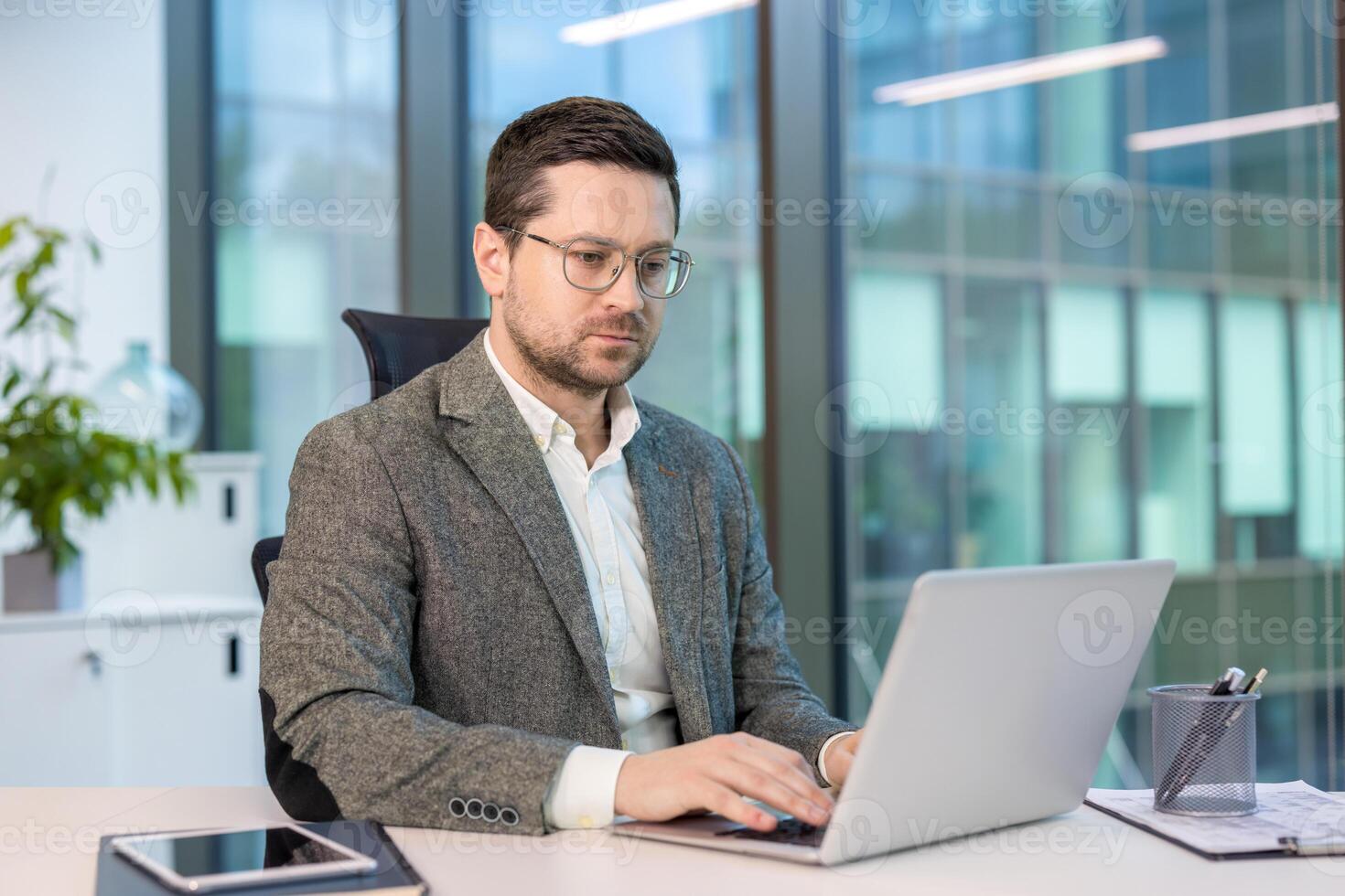 A serious young businessman in a suit is sitting at a desk in the office and working intently on a laptop. photo