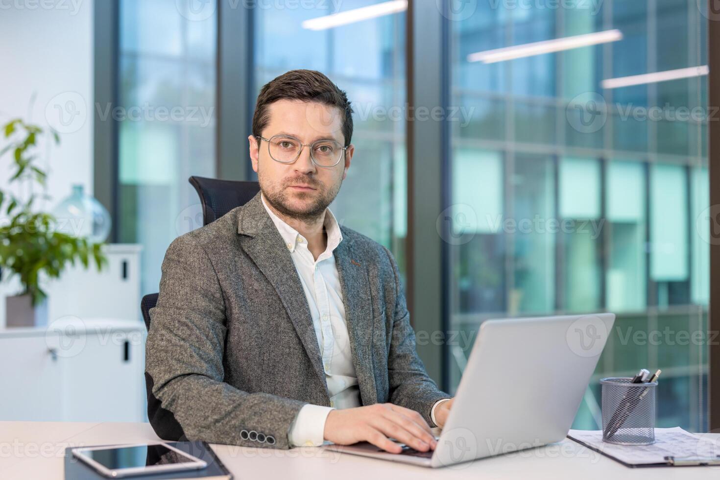 Portrait of a serious and confident young man in a suit and glasses sitting in the office at a desk, working on a laptop and looking at the camera. photo
