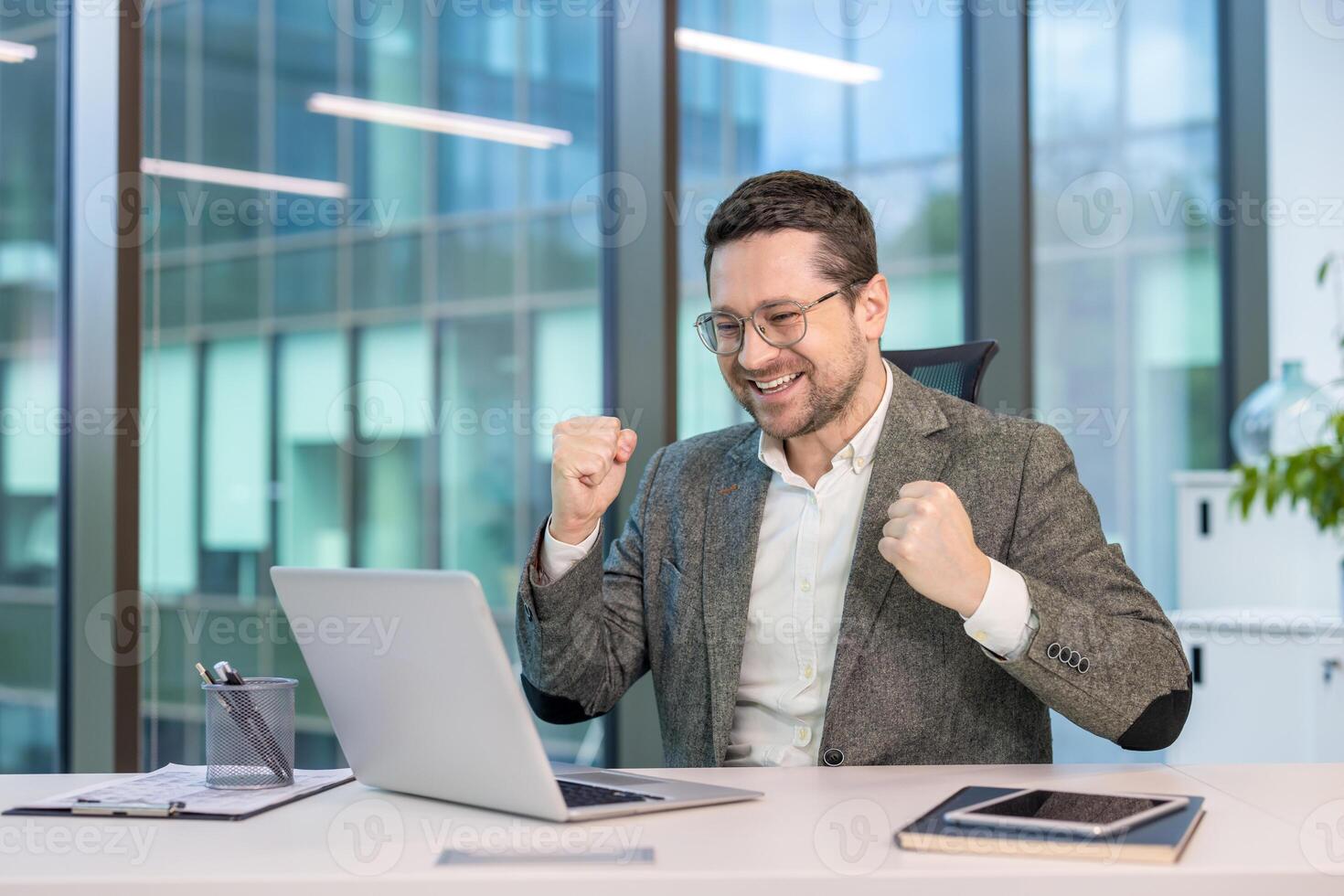 Happy young businessman sitting in the office at his desk, looking at the laptop screen and rejoicing, showing a gesture of victory and success. photo