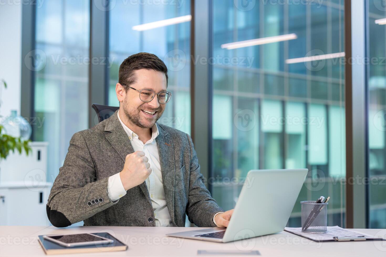 Happy young businessman sitting at a desk in the office, looking at the laptop screen and rejoicing at the news and smiling, showing a victory gesture with his hand. photo