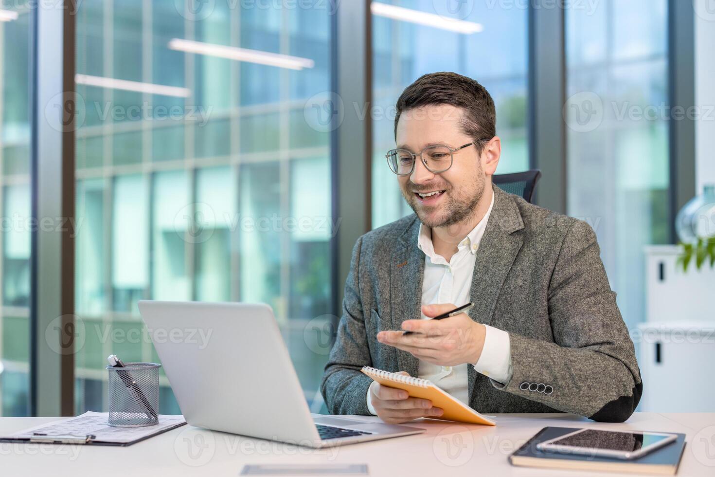 Smiling young businessman in a suit sitting at his workplace in the office and talking remotely via laptop, making notes in a notebook. photo