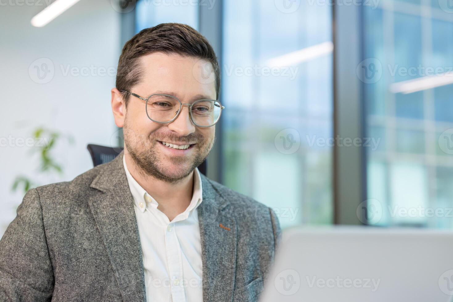 Smiling young male businessman sitting in the office at a desk, working on a laptop and talking on a call with clients. photo