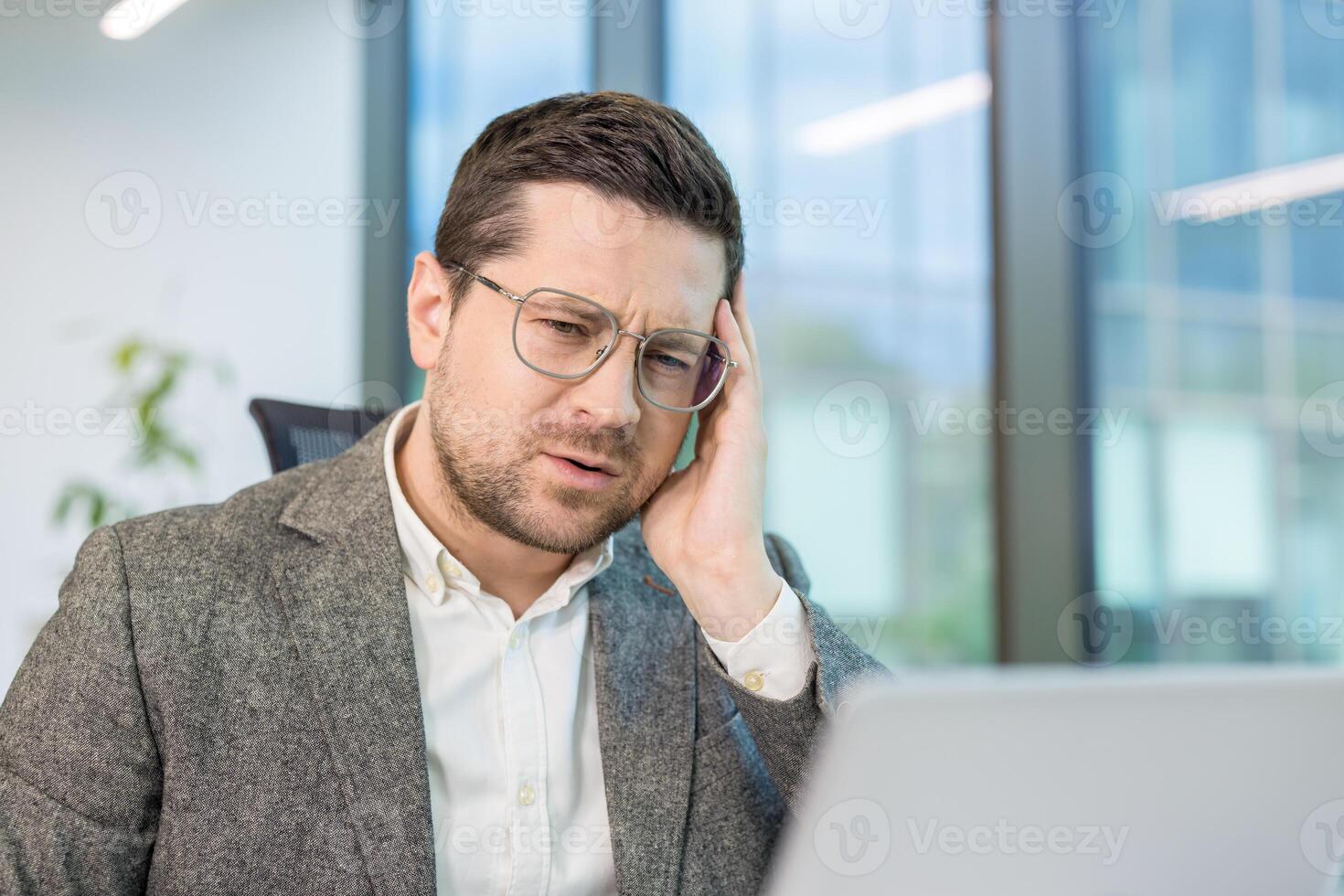 Close-up portrait of a sad and worried young man holding his head and looking at the laptop screen at his workplace in the office. photo