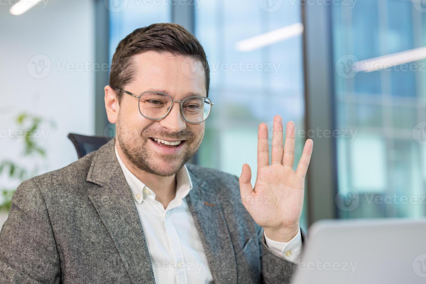 A smiling businessman waves during a call, working in a modern office environment. photo