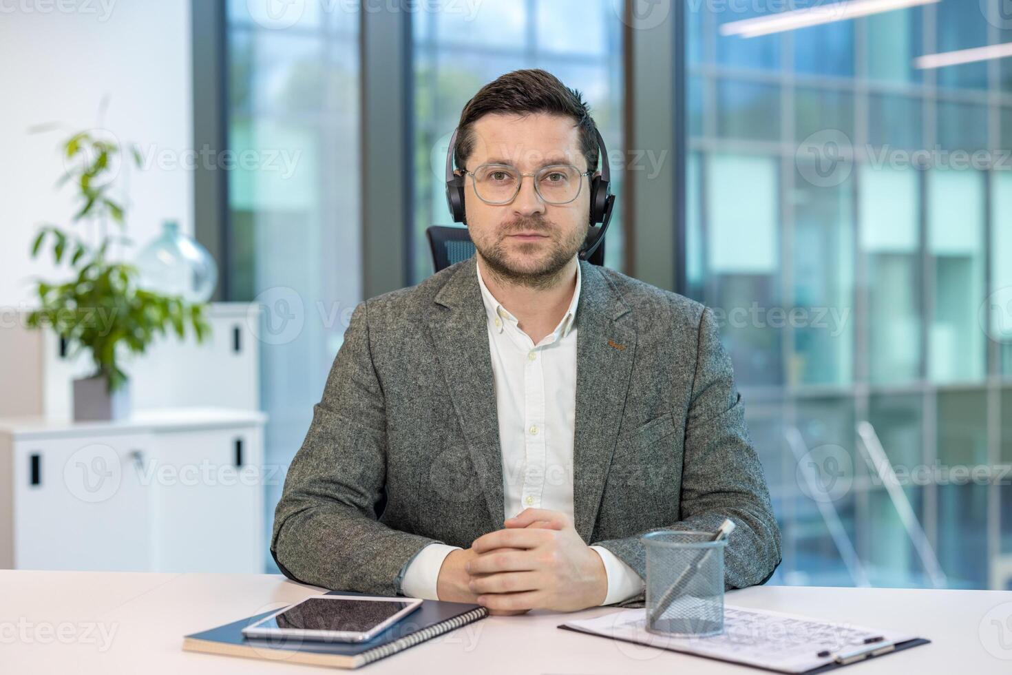 Close-up portrait of a smiling young man in a headset and suit sitting at a desk in the office, talking and waving at the camera. photo