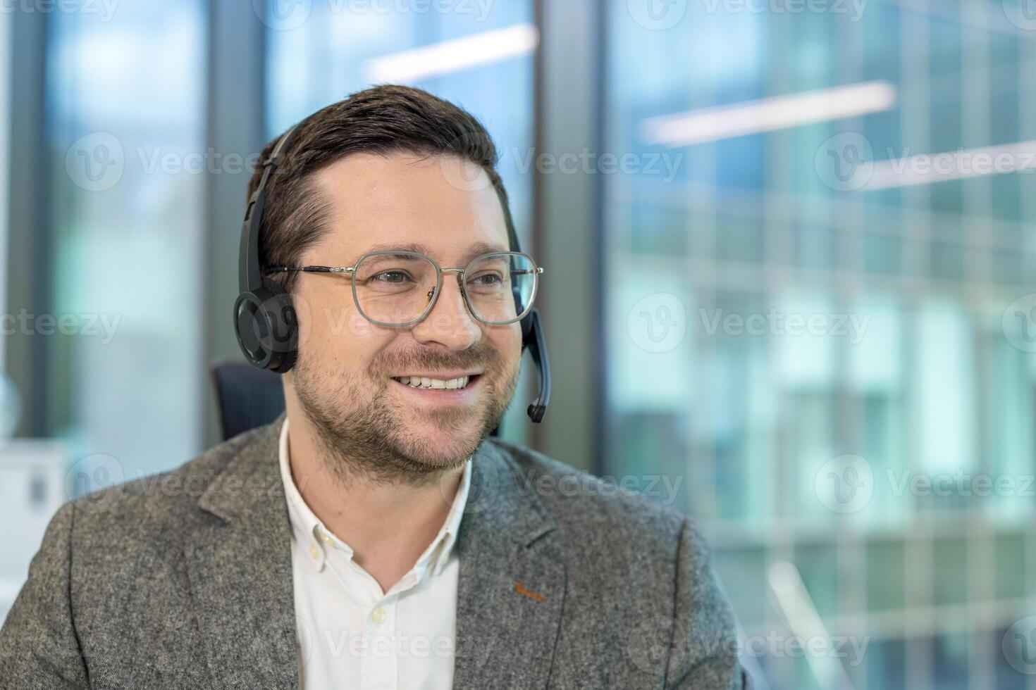 A close-up photo of a young man wearing a headset and glasses who is working in an office, smiling and looking to the side.