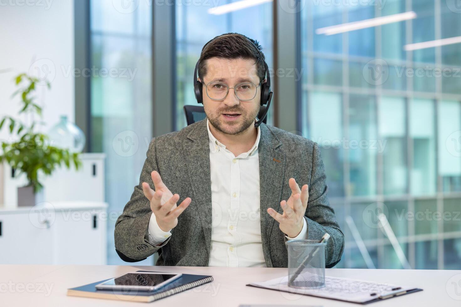 Portrait of a young man in a headset and computer sitting in an office at a table and holding an online conference, explaining by gesturing with his hands. photo