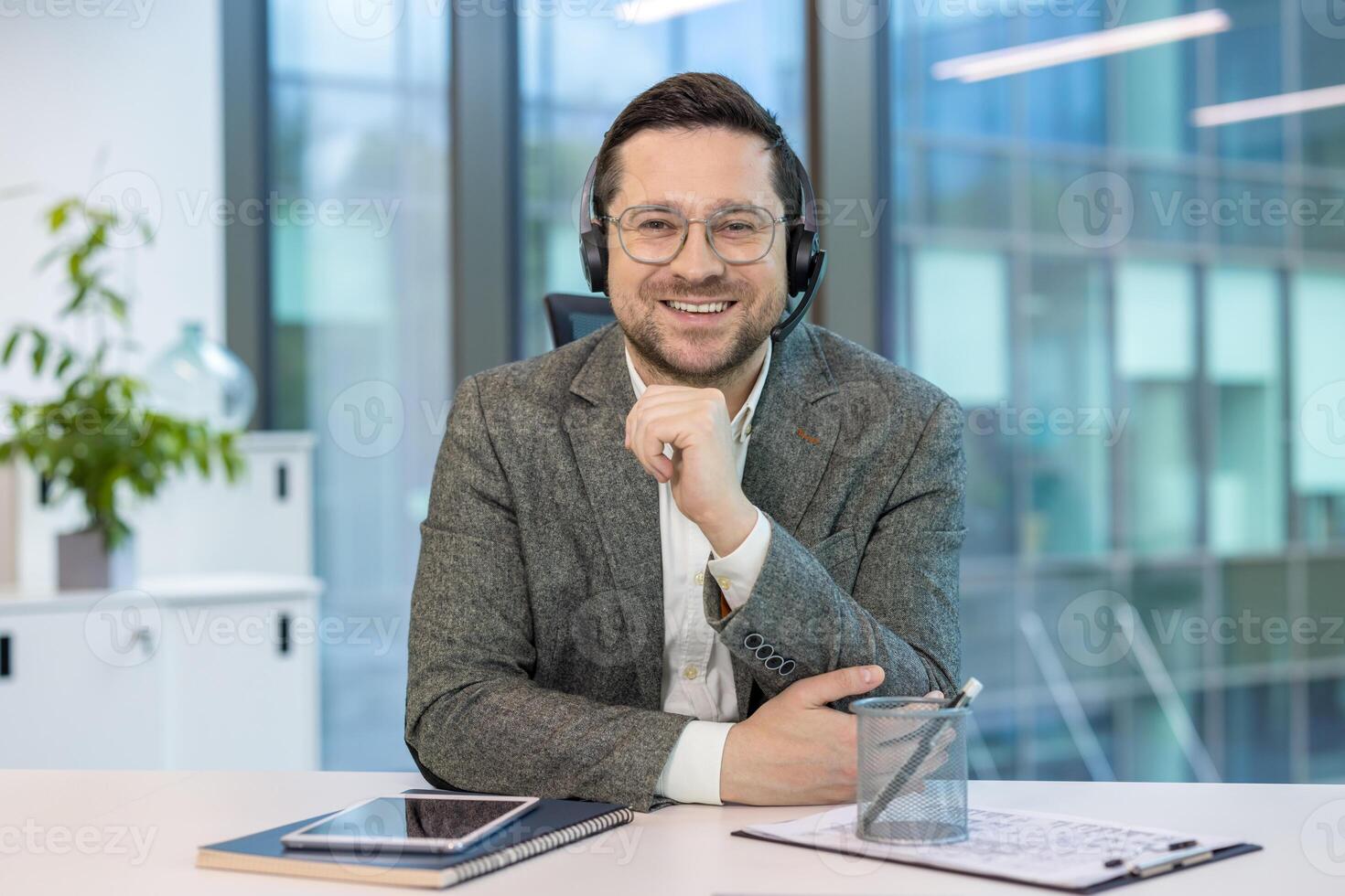 Portrait of a smiling young businessman in a headset and suit sitting at a desk in the office, looking and smiling confidently at the camera. photo