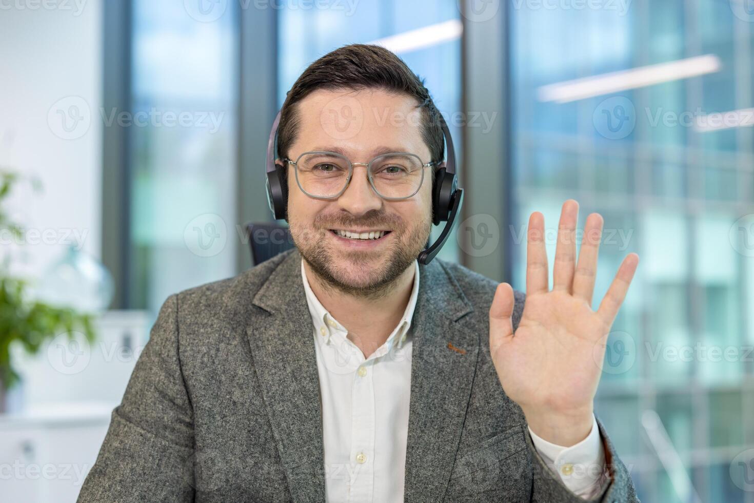 Close-up portrait of a young man in a suit and headset sitting at a desk in an office, looking and waving at the camera. photo