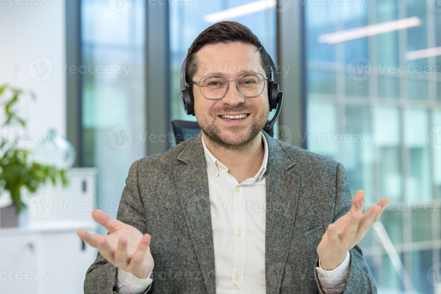 Portrait of a smiling young man in a headset sitting at an office desk and talking to the camera while gesturing with his hands. photo