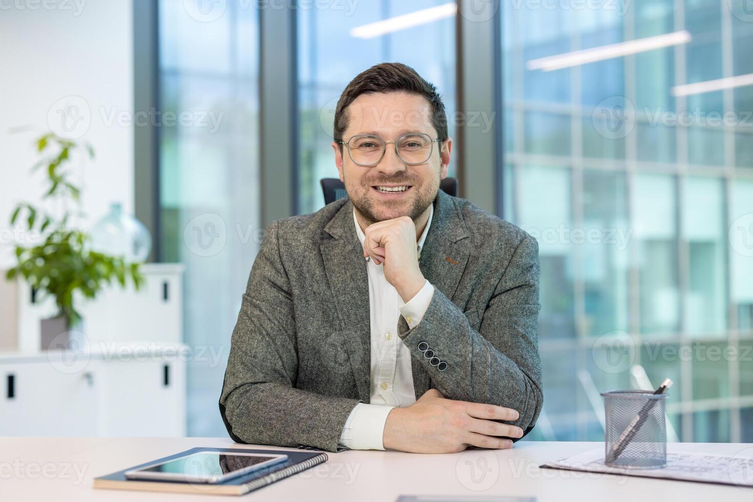 Portrait of a young businessman sitting in the office at a desk in a suit and glasses, smiling and looking at the camera. photo
