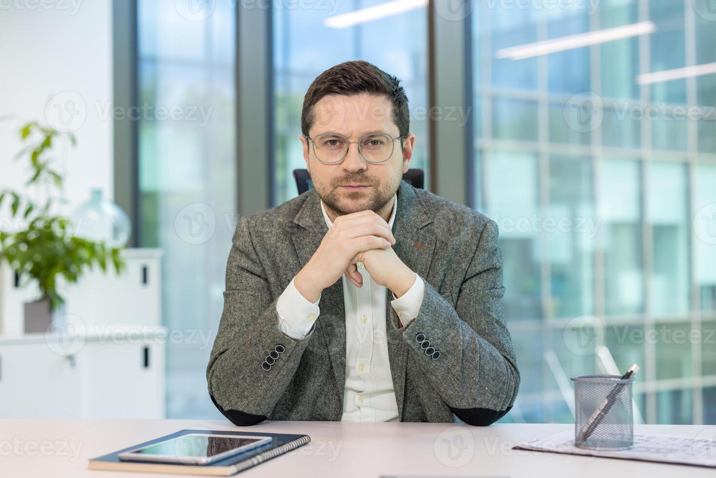 Portrait of a young successful and serious man in a suit and glasses sitting at a desk in the office, and looking confidently at the camera. photo