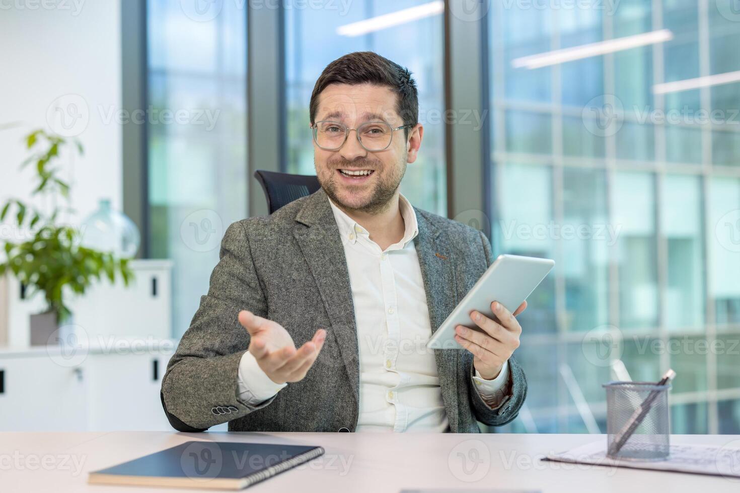 A businessman in a conference, holding a tablet and gesturing in an office setting. He appears to be in a virtual meeting photo