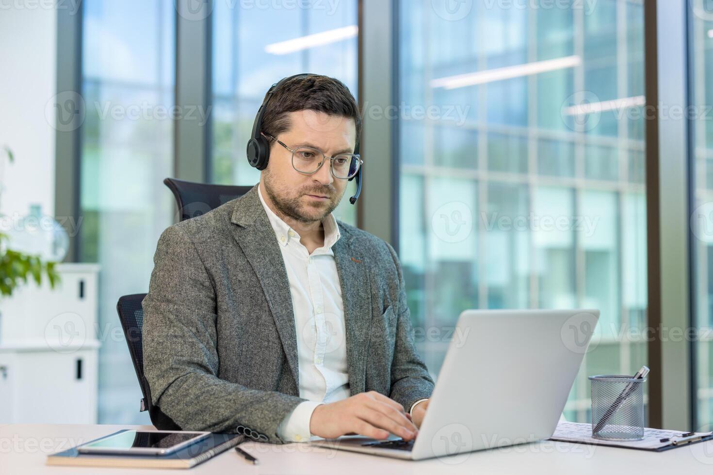 Serious young man in a suit and headset working intently on a laptop while sitting at a desk in the office. photo
