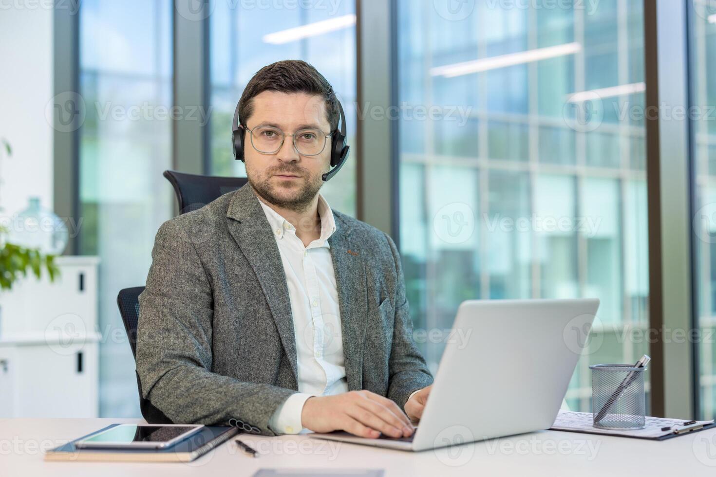 Serious young man, office worker and consultant sitting at desk, working on laptop and looking at camera. photo