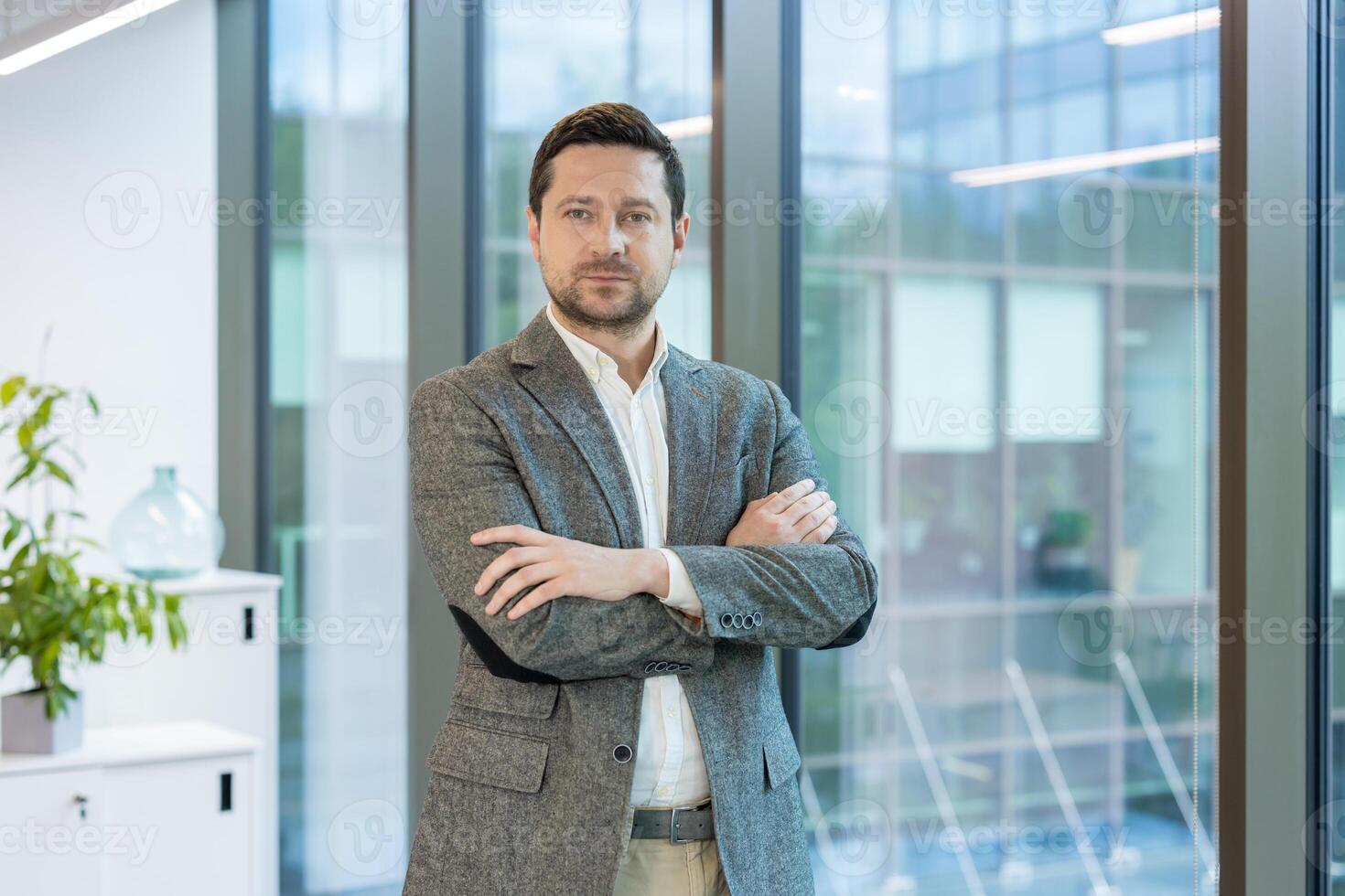 Young successful businessman standing in the office in a suit and tie, arms crossed on his chest, looking confidently at the camera. photo