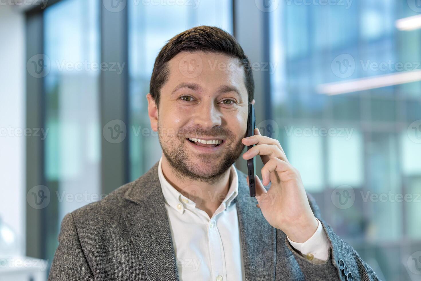 Close-up photo of a young male office worker standing inside a building, talking on the phone and looking at the camera with a smile.