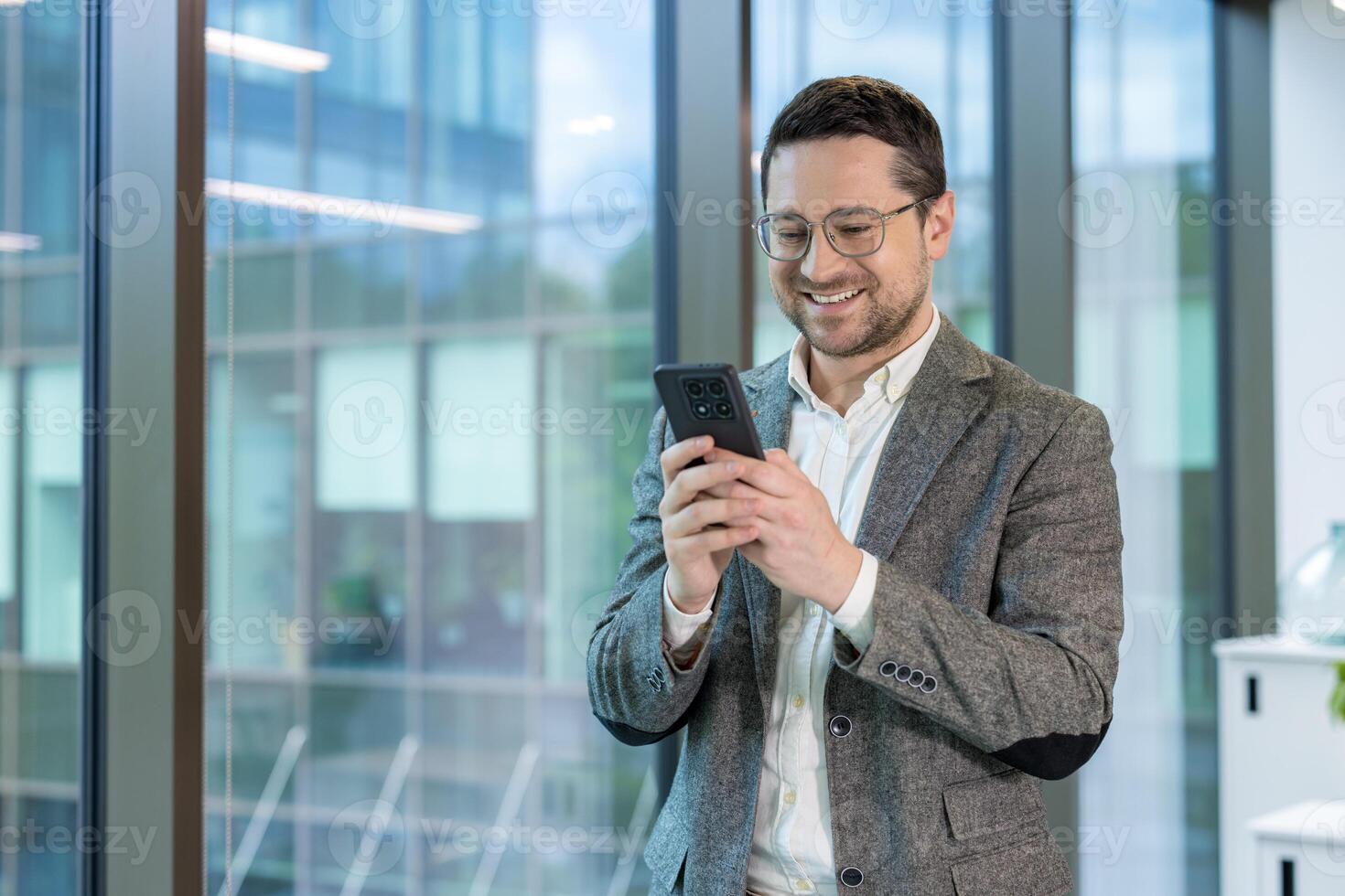 A smiling businessman wearing glasses, using a smartphone, standing near a large window in a modern office setting. photo