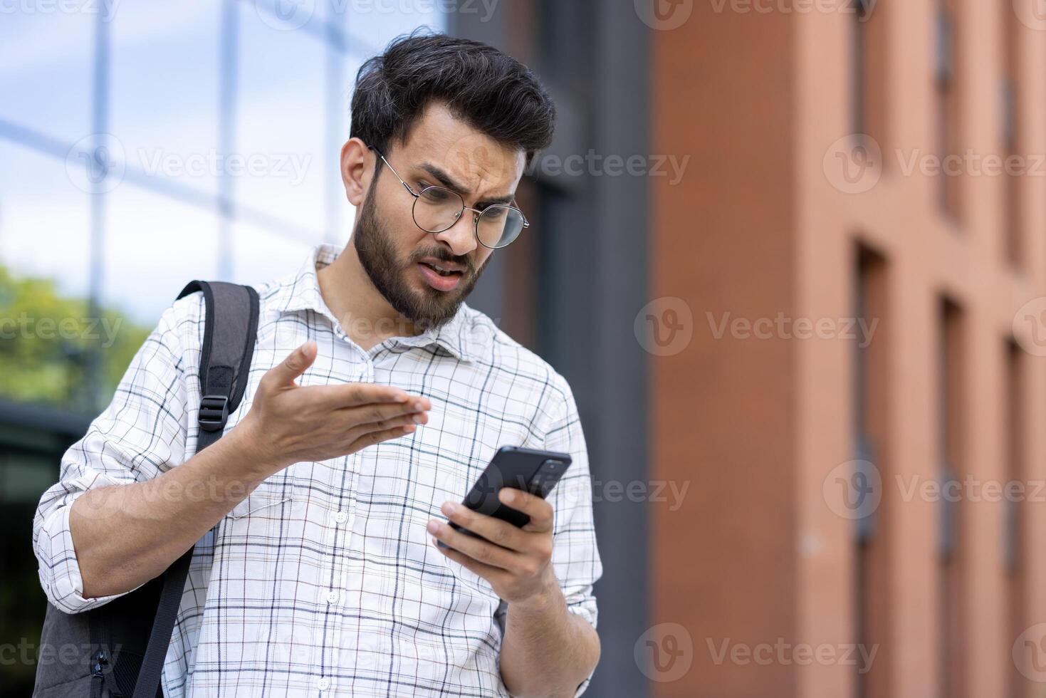 A young male student wearing glasses, looking confused at his phone while standing outside a building. Possibly dealing with a problem. photo