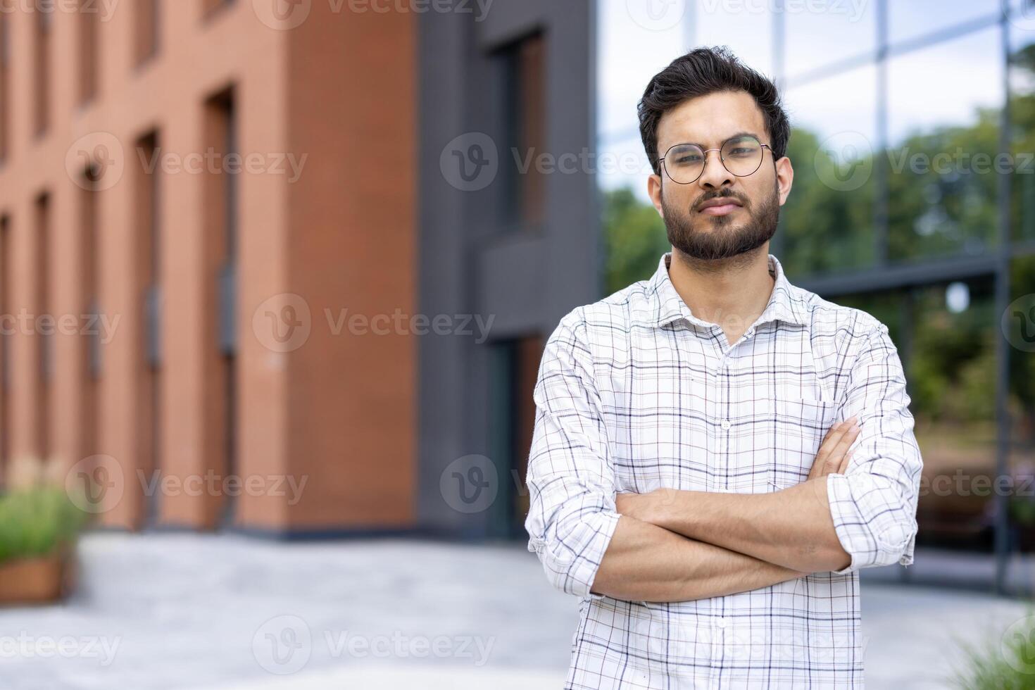An Indian man with a serious expression stands in front of a modern building with his arms crossed. photo