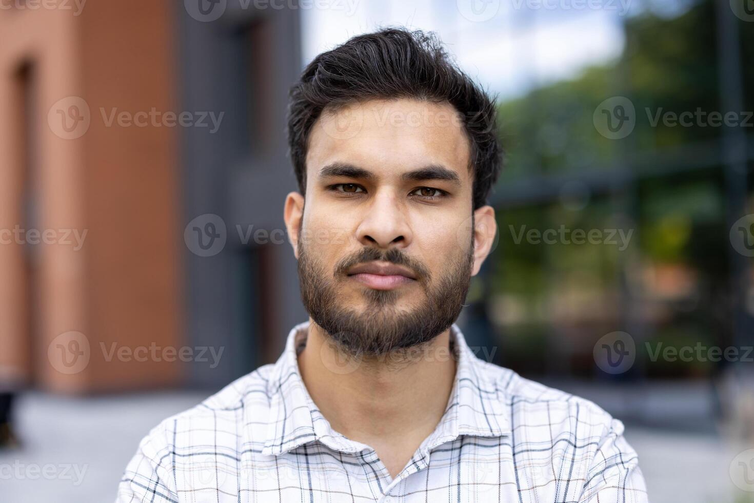 A focused Indian man gazes directly at the camera, captured in a close-up shot. He is wearing a checkered shirt outside. photo