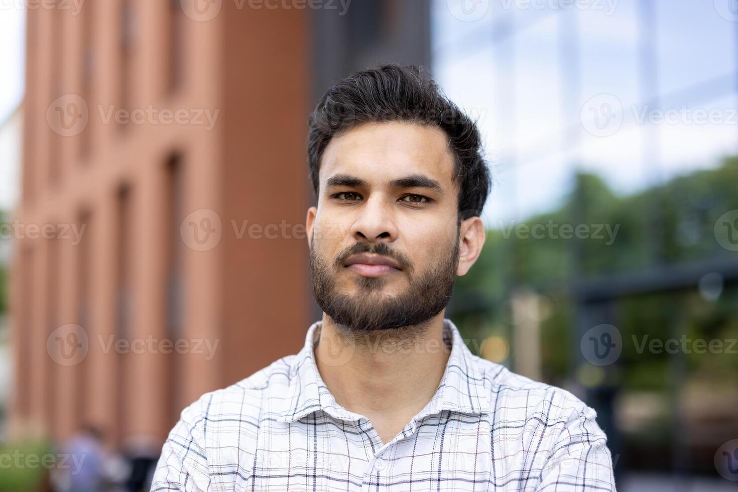Close-up portrait of a young Muslim man in a shirt standing outside an office building, university campus, looking seriously at the camera. photo